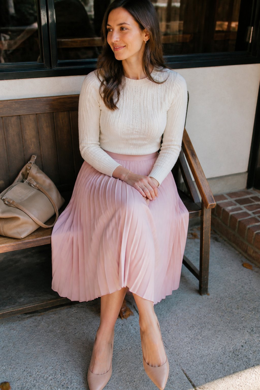 A woman wearing a pastel pleated midi skirt and fitted knit sweater standing near a wooden bench with a handbag nearby, lit by soft natural daylight.
