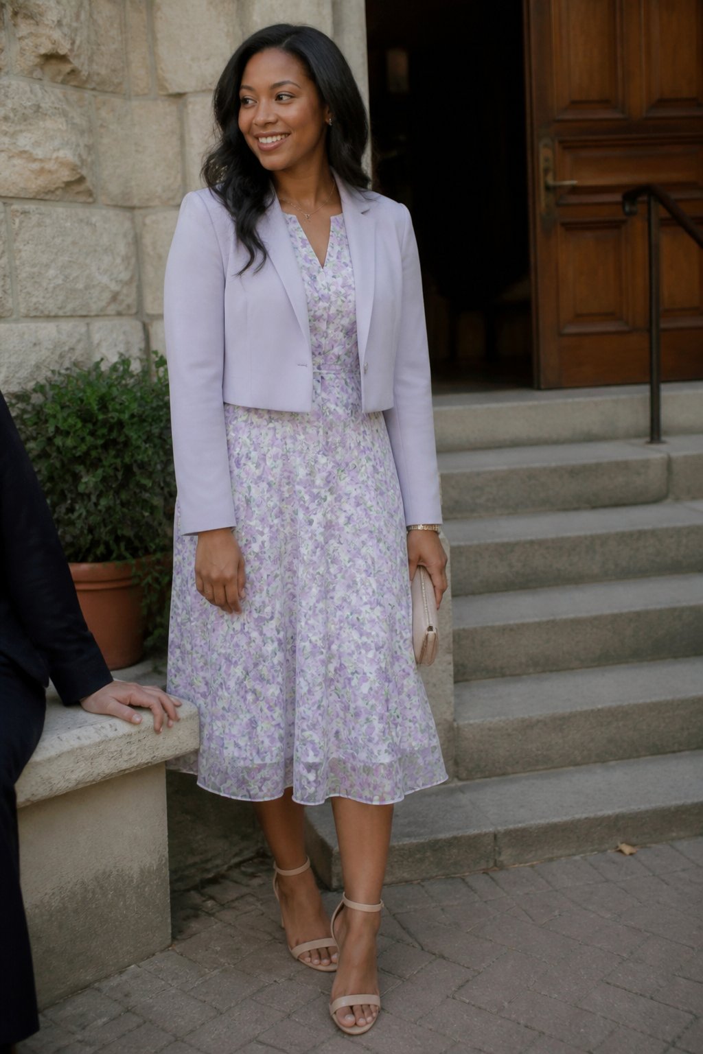 A woman wearing a cropped blazer over a floral tea dress and ankle-strap heels, standing indoors with soft daylight coming through a window.