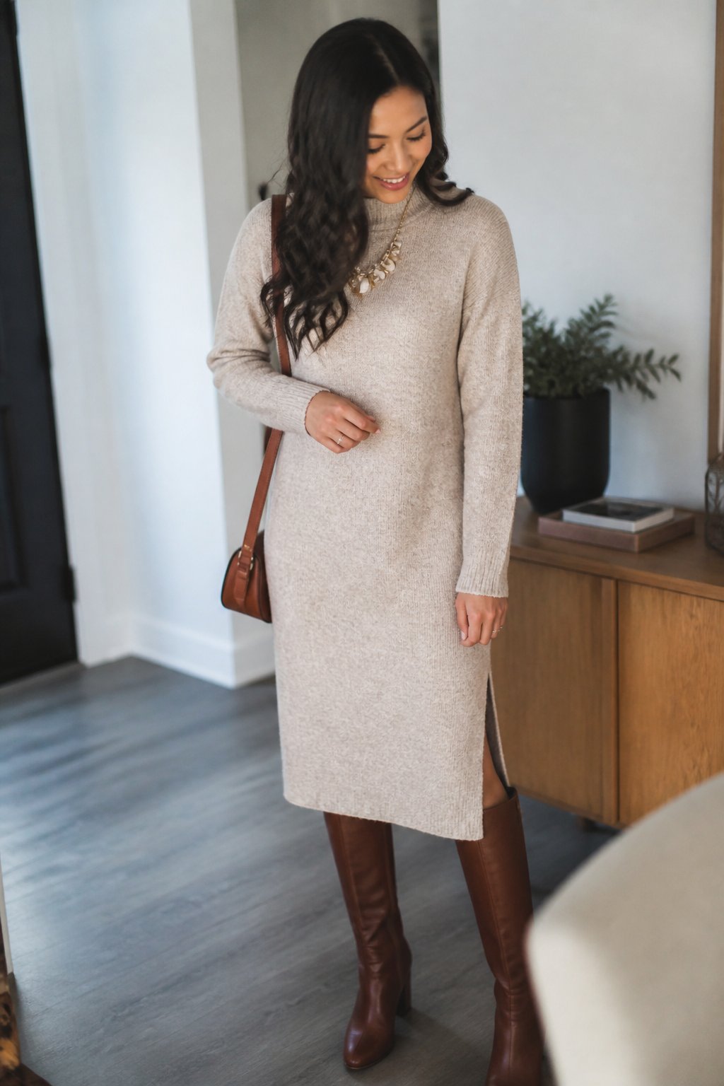 A woman wearing a soft knit midi sweater dress, statement necklace, and knee-high boots standing indoors with natural daylight.
