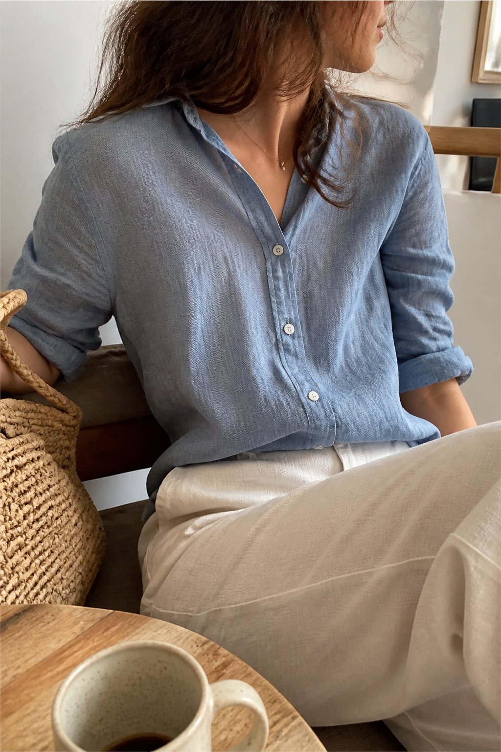 A lightweight linen button-up shirt shown casually in a summer setting with soft daylight, surrounded by everyday objects like a wooden chair and a ceramic mug.