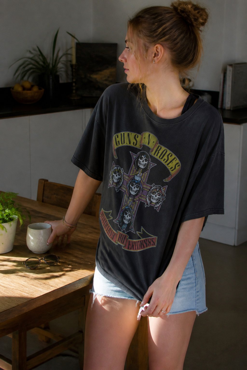 A person wearing an oversized graphic band tee standing near a wooden table with sunglasses, a mug, and a small plant in soft natural daylight.