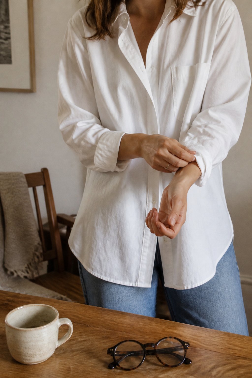 A person adjusting a white button-down shirt in a cozy indoor setting with natural daylight and everyday objects around.