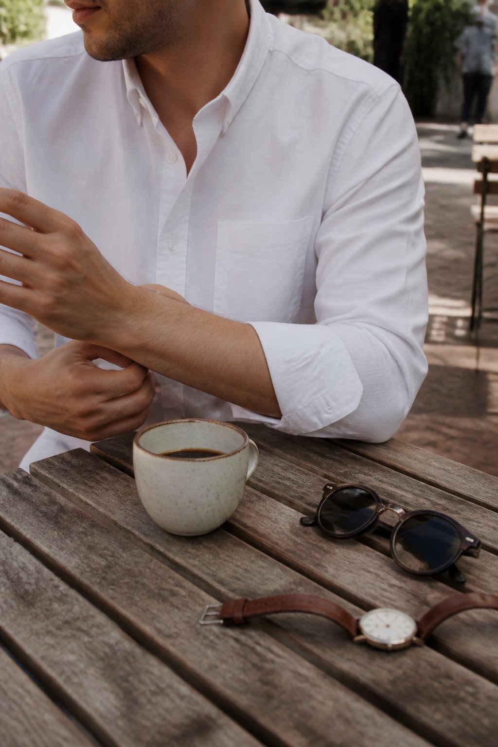 Person wearing a white button-down shirt in a casual setting with natural light, sitting at a wooden table with a coffee cup and sunglasses nearby.