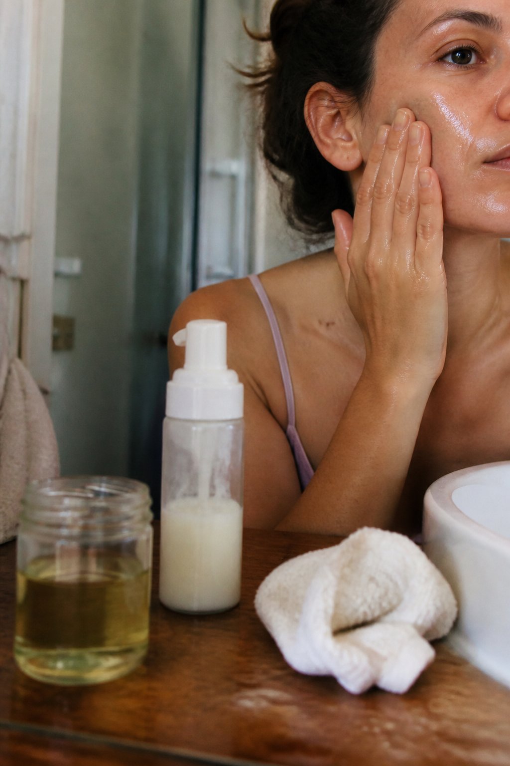 Close-up of hands applying oil-based cleanser on skin next to foaming cleanser bottle and cotton washcloth on a bathroom countertop.