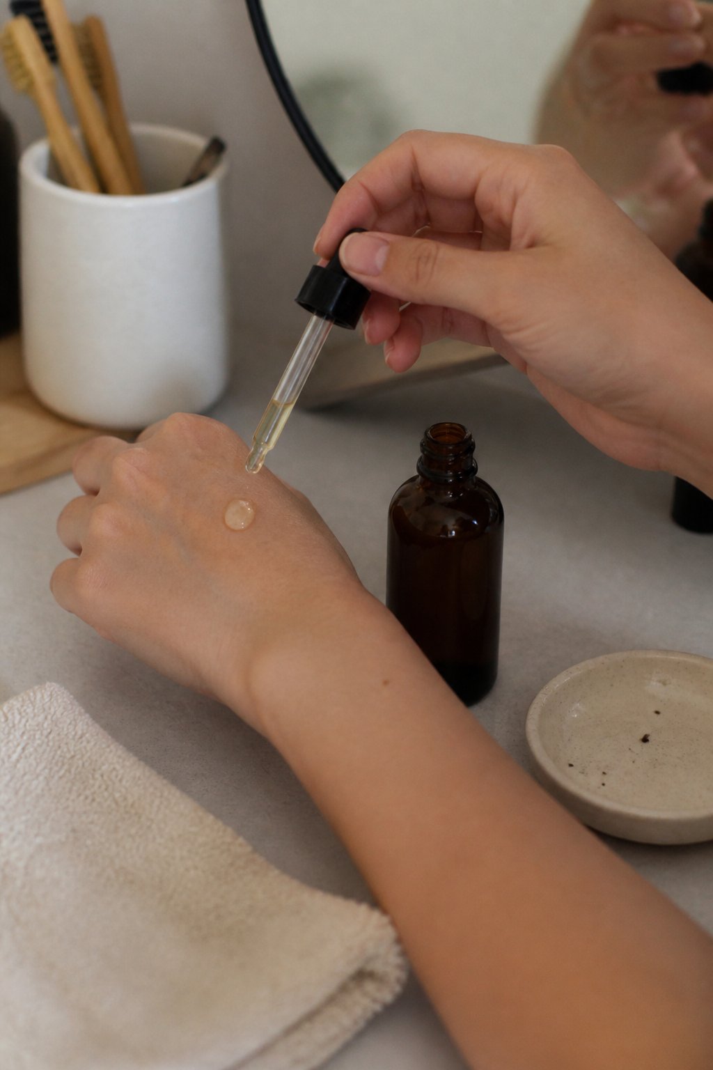 Close-up of a person applying vitamin C serum to their hand on a bathroom counter with natural daylight and everyday skincare items nearby.