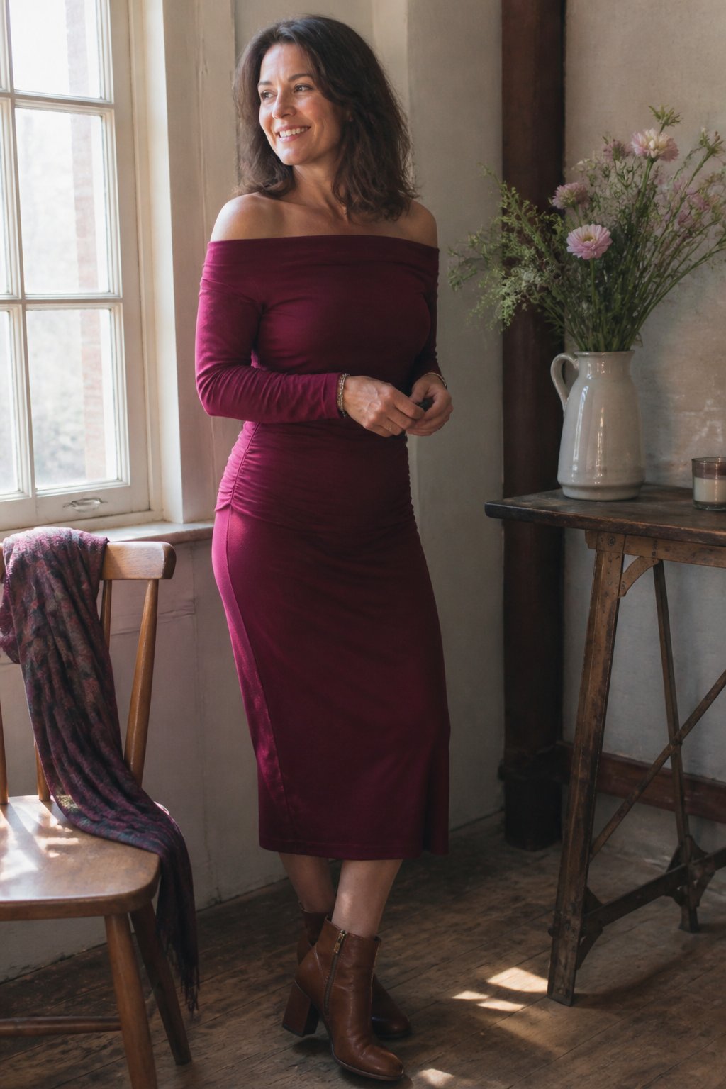 A woman over 40 wearing a burgundy off-shoulder midi dress and block-heel ankle boots stands near a window with natural light, surrounded by a wooden chair and a vase with flowers.