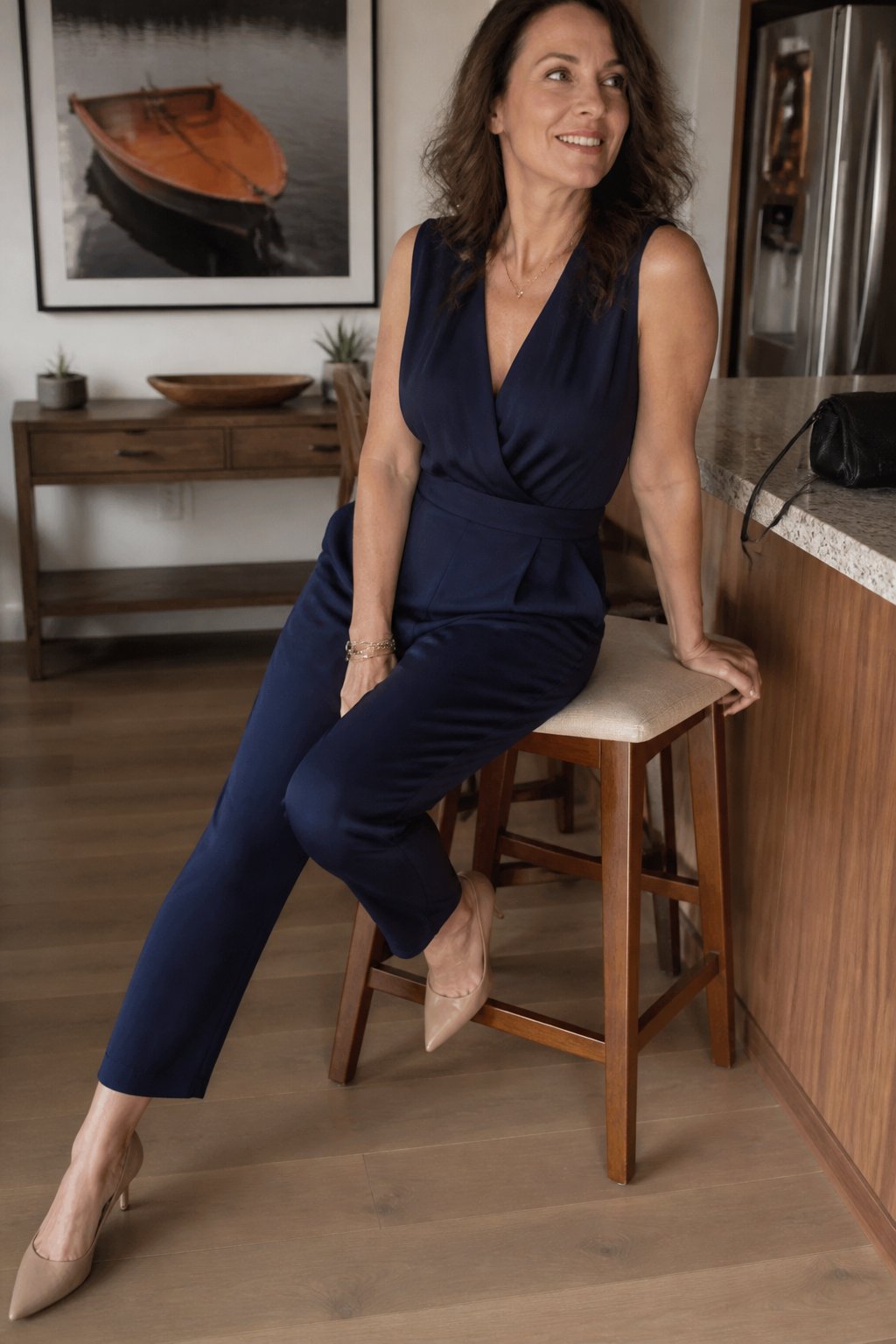 A woman over 40 wearing a navy V-neck jumpsuit and pointed-toe pumps in a softly lit indoor setting.