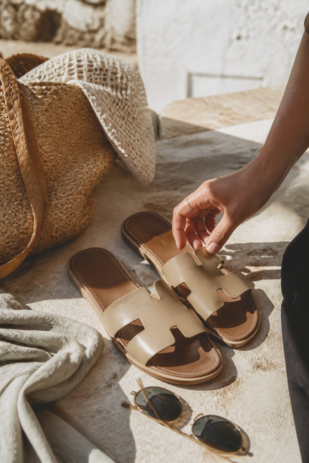 A pair of neutral leather slide sandals on a sunlit surface with a hand adjusting one sandal, surrounded by summer accessories like a woven bag and sunglasses.