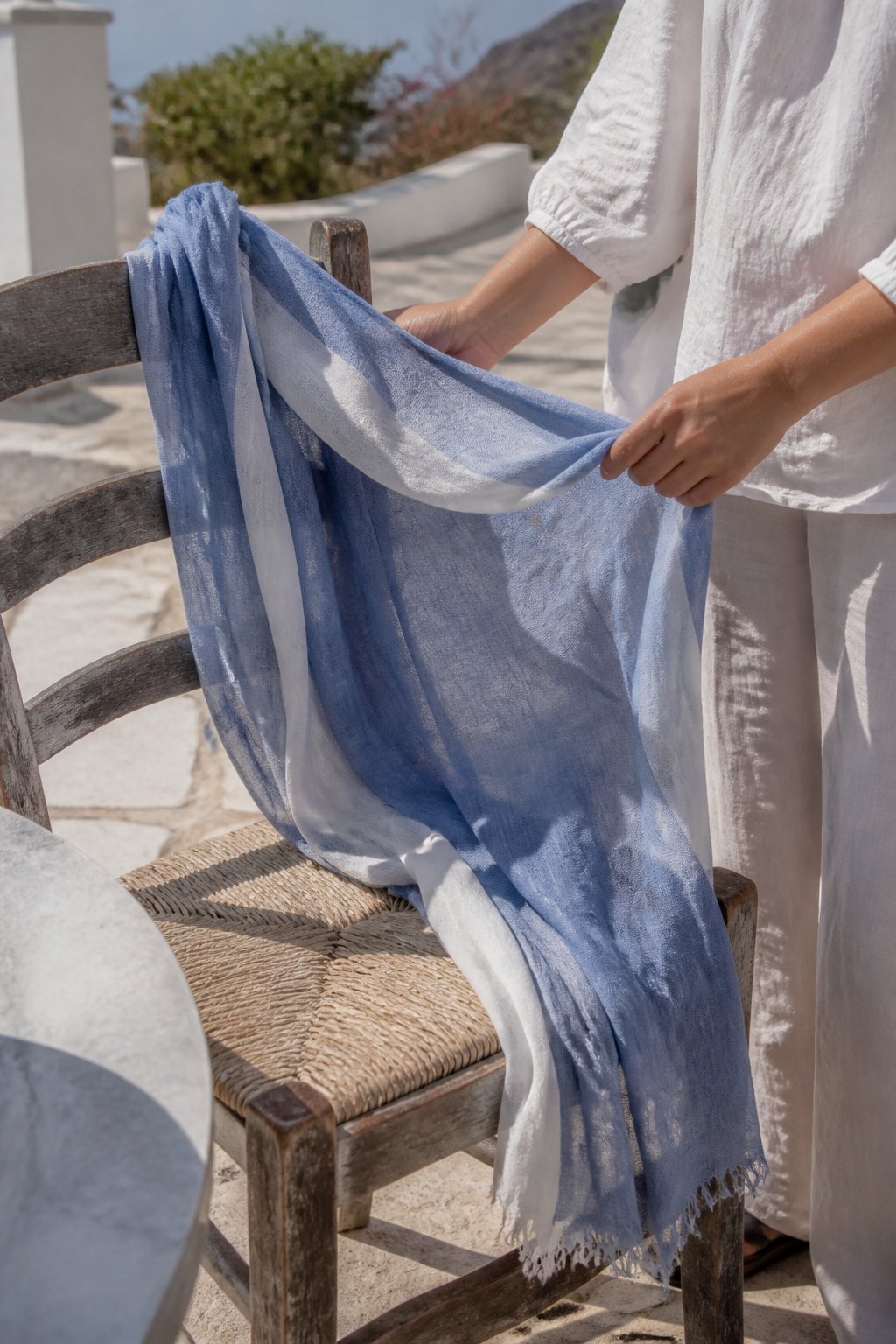 A lightweight scarf draped casually outdoors on a wooden chair in soft daylight with a blurred background of Mediterranean greenery.