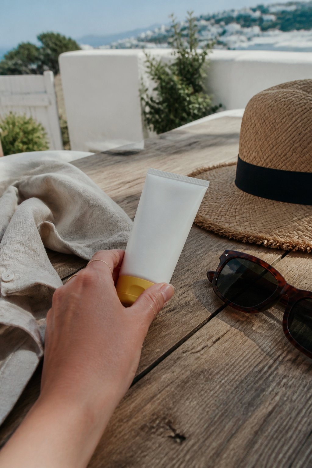 A hand reaching for a bottle of reef-safe sunscreen on a wooden table with a linen shirt, sunglasses, and a straw hat, set on a sunlit terrace with white walls and greenery in the background.