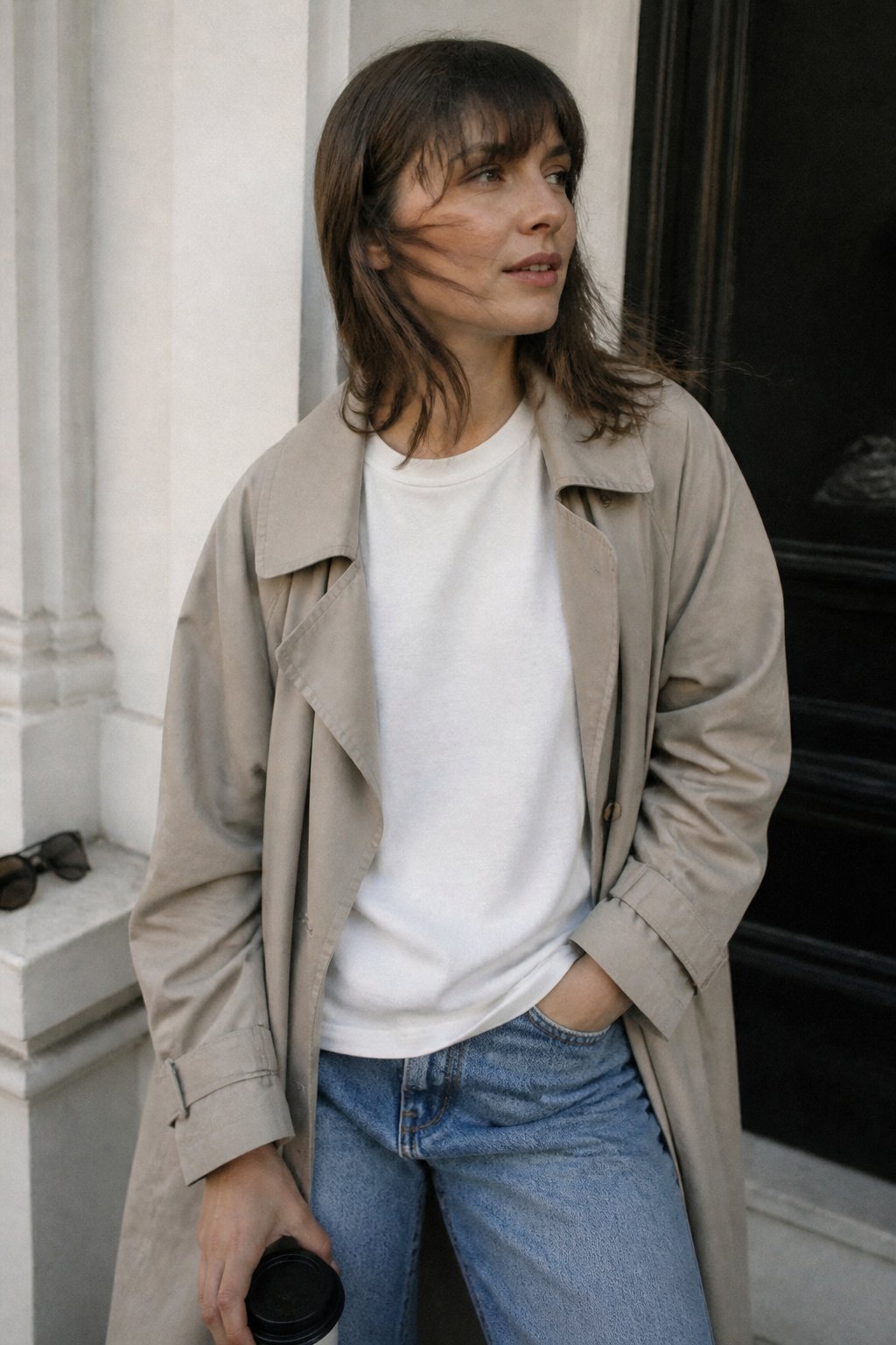 A person wearing a lightweight trench coat layered over a cotton tee and denim jeans, standing casually in soft daylight with natural shadows and a slightly off-center composition.
