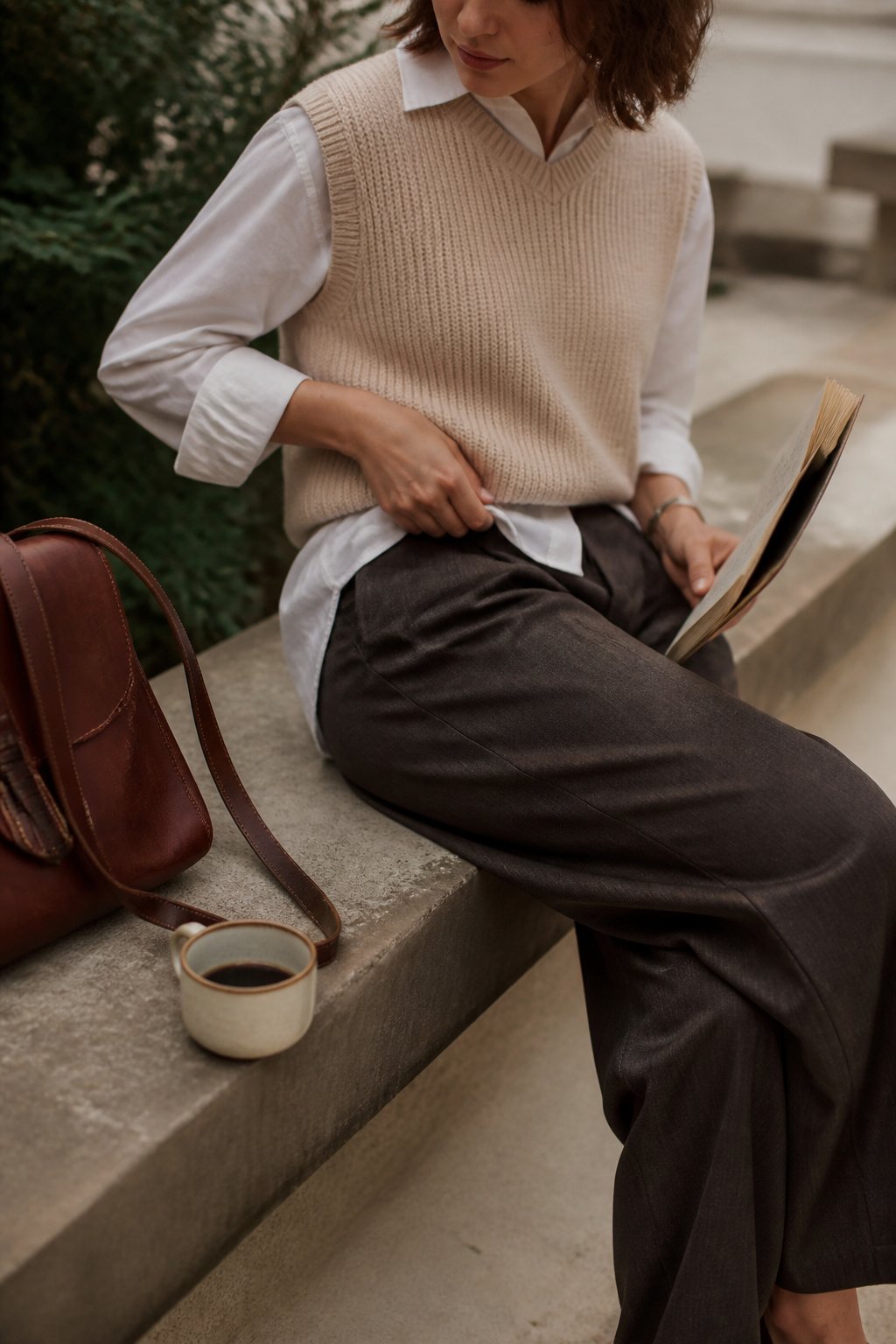 Person wearing a sleeveless knit vest over a long-sleeve button-down shirt and wide-leg trousers, standing casually with hands adjusting the vest, surrounded by everyday objects like a leather bag and coffee cup.