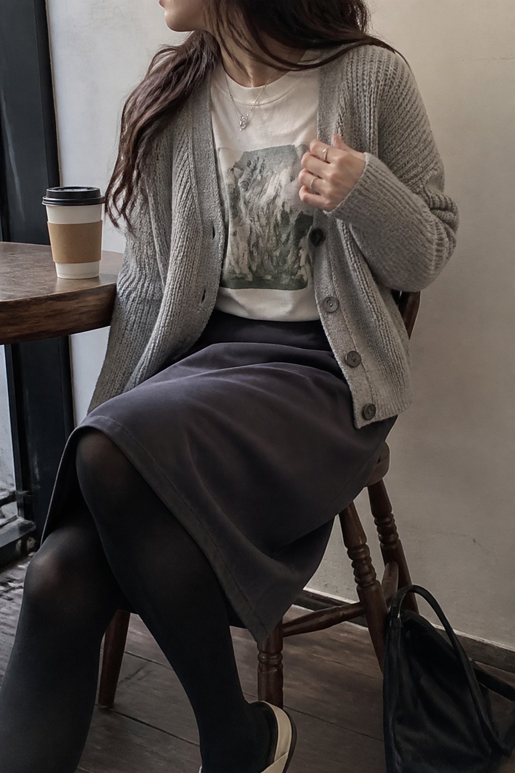 Person wearing an oversized cardigan, graphic tee, mid-length skirt, and opaque tights standing indoors near a window with soft daylight, with a coffee cup and a bag nearby.