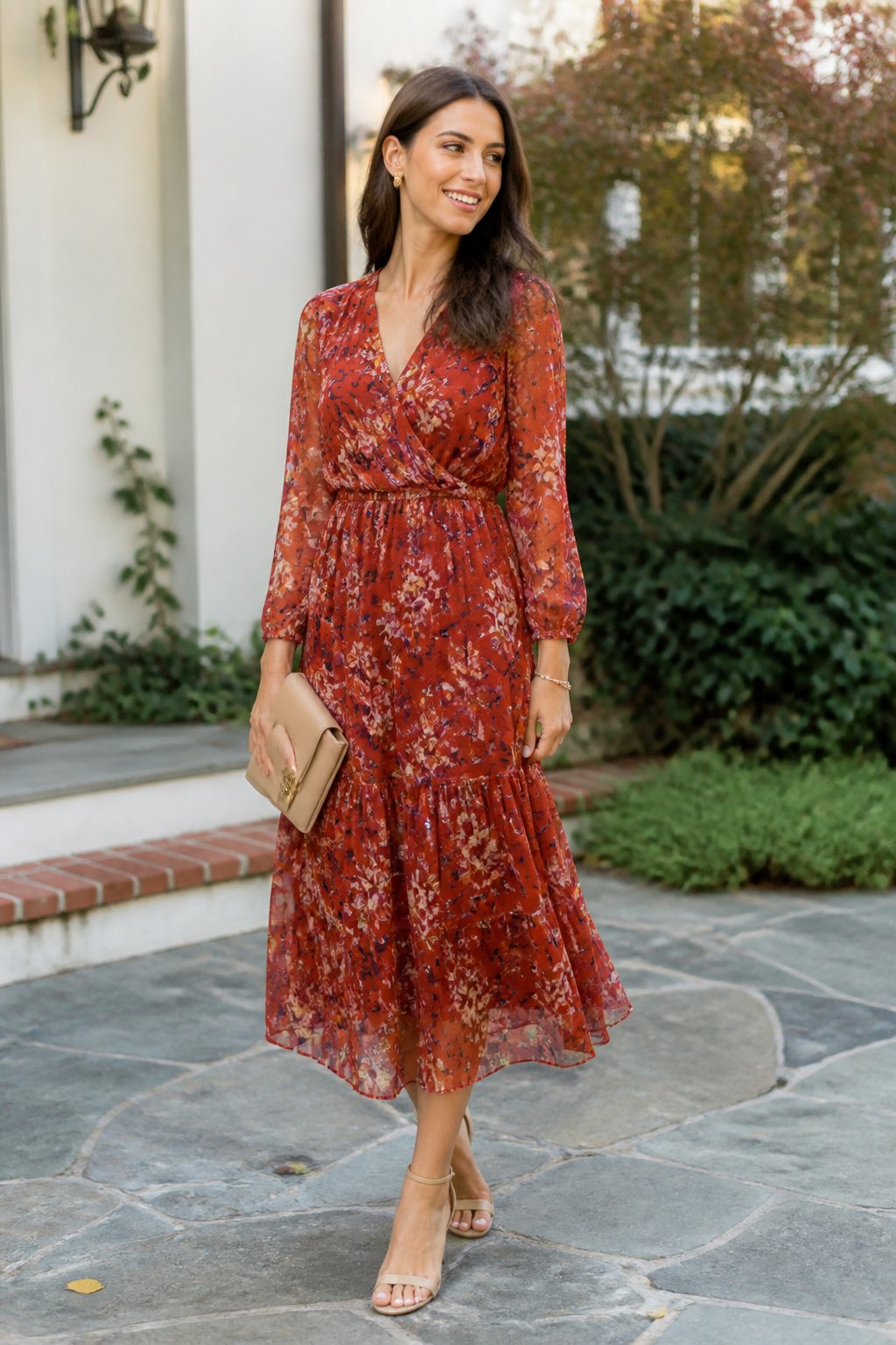 A woman wearing a rust floral-print chiffon midi dress stands outdoors among soft autumn foliage.