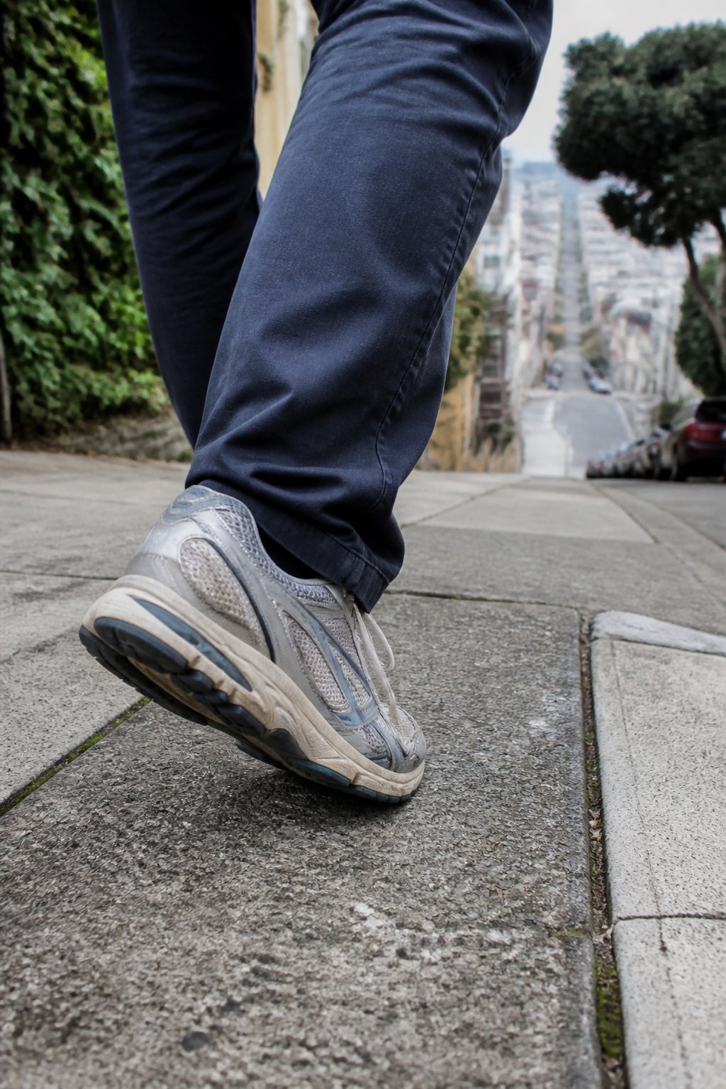 A person wearing supportive walking sneakers steps along a gently sloped city sidewalk.