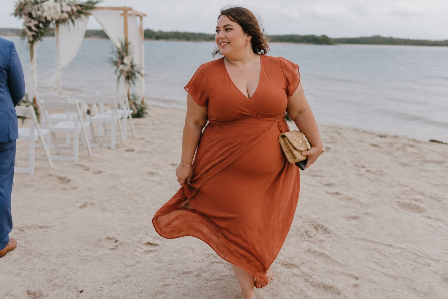 A plus-size woman in an elegant flowing dress stands near the ocean at a beach wedding.