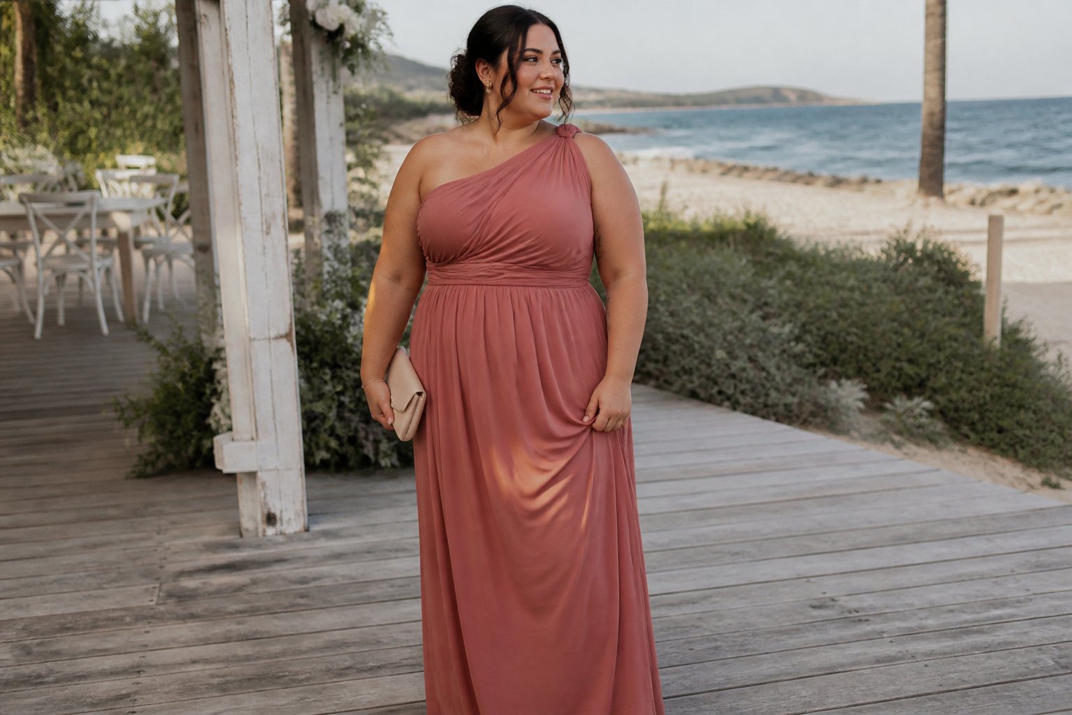 A plus-size woman in a one-shoulder maxi dress standing near a beach wedding setting.
