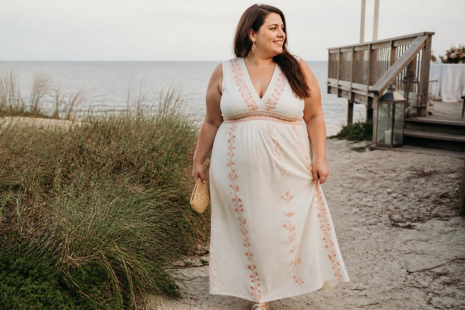 A plus-size woman wearing an embroidered cotton maxi dress walks beside a beach with the ocean in the background.