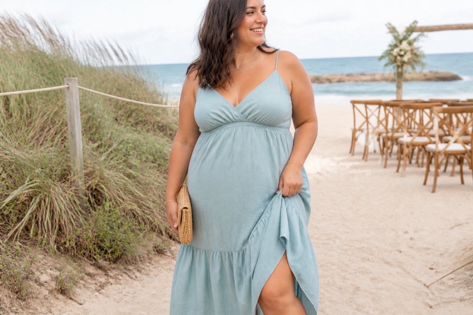 A plus-size woman wearing a flowing spaghetti-strap empire-waist dress stands on a sandy beach near dune grass and a simple wedding setup.