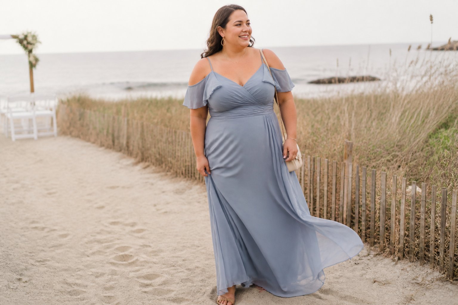 A plus-size woman in a flowing cold-shoulder chiffon maxi dress standing near a sandy beach with dune grass.