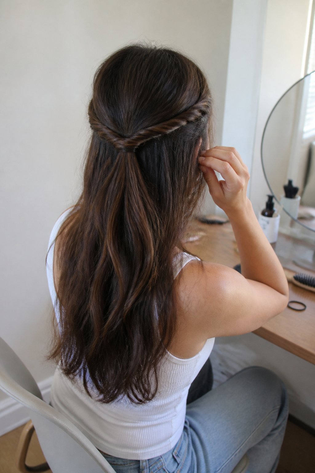 A woman with long hair wears a relaxed half-up rope twist while standing near a bedroom or bathroom vanity.