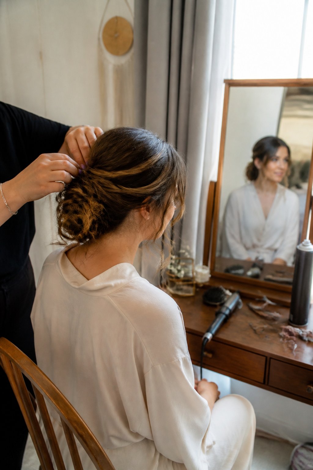 A hairstylist arranges a guest’s softly pinned hairstyle beside a mirror, with styling tools nearby.