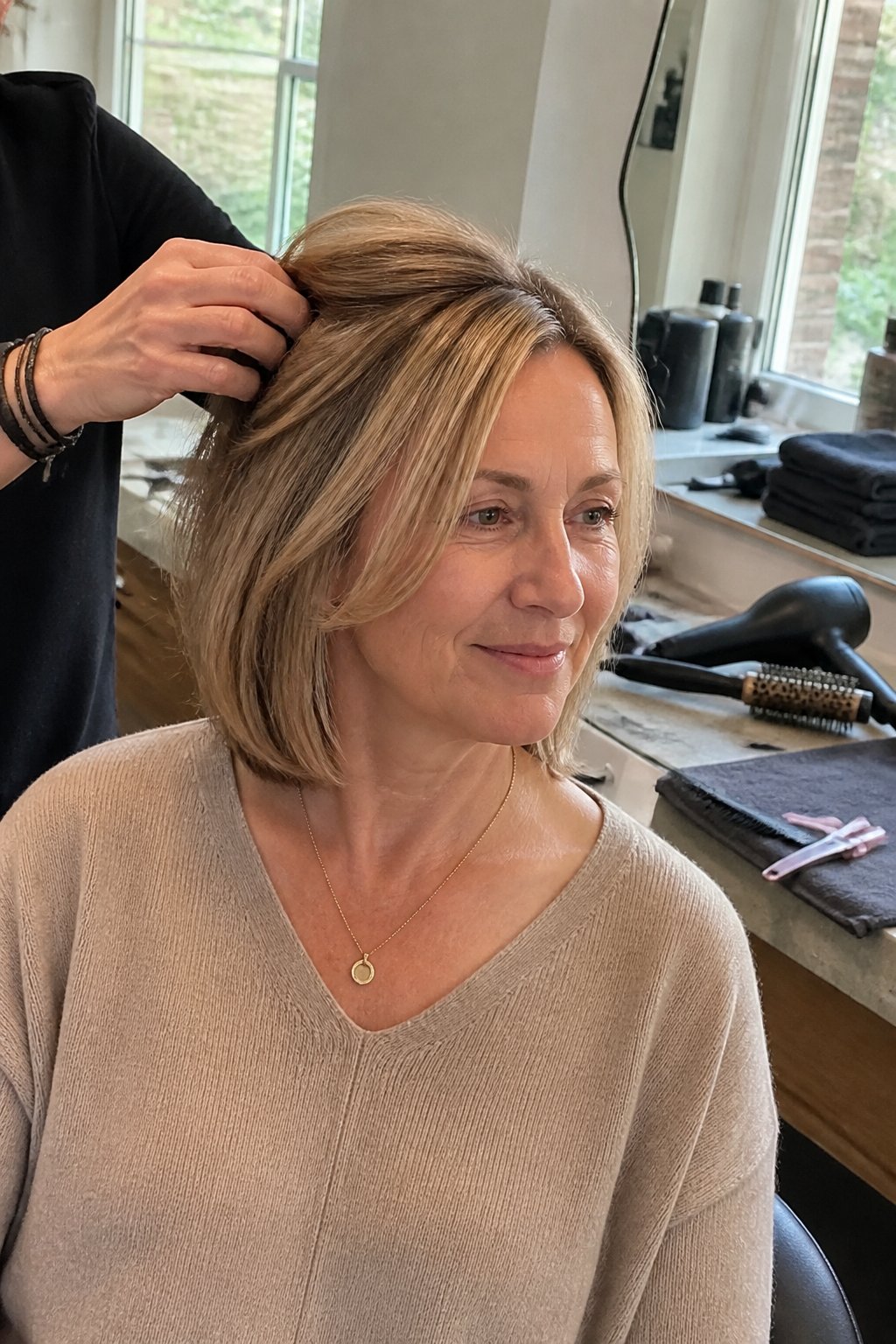 A hairstylist shapes a woman’s fine hair in a bright salon using a comb and styling tools.