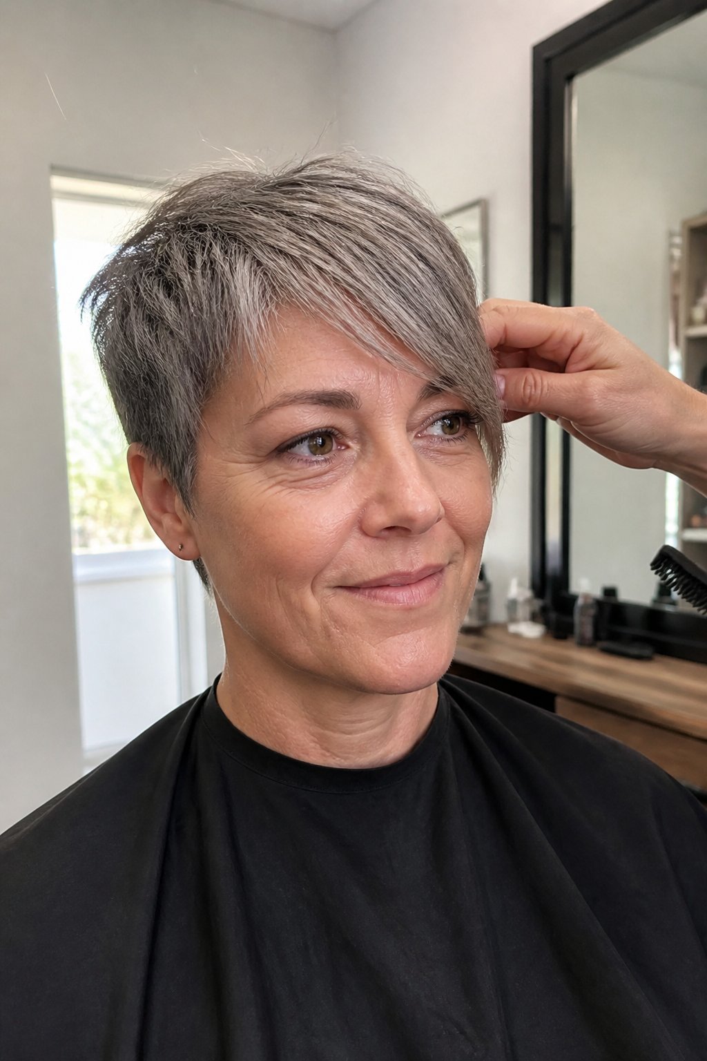 A woman over 40 with an asymmetrical pixie haircut and a longer sweeping fringe poses in a softly lit everyday interior.