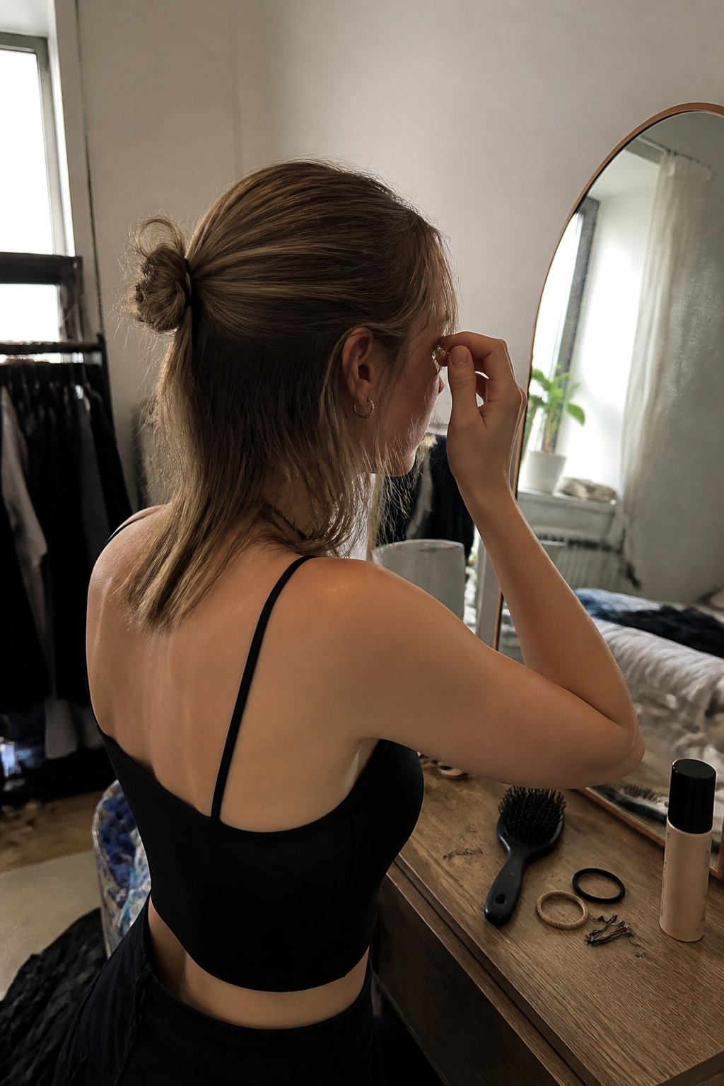 A person with thin hair adjusts a simple concert-ready hairstyle beside a mirror, with hair tools on a nearby surface.