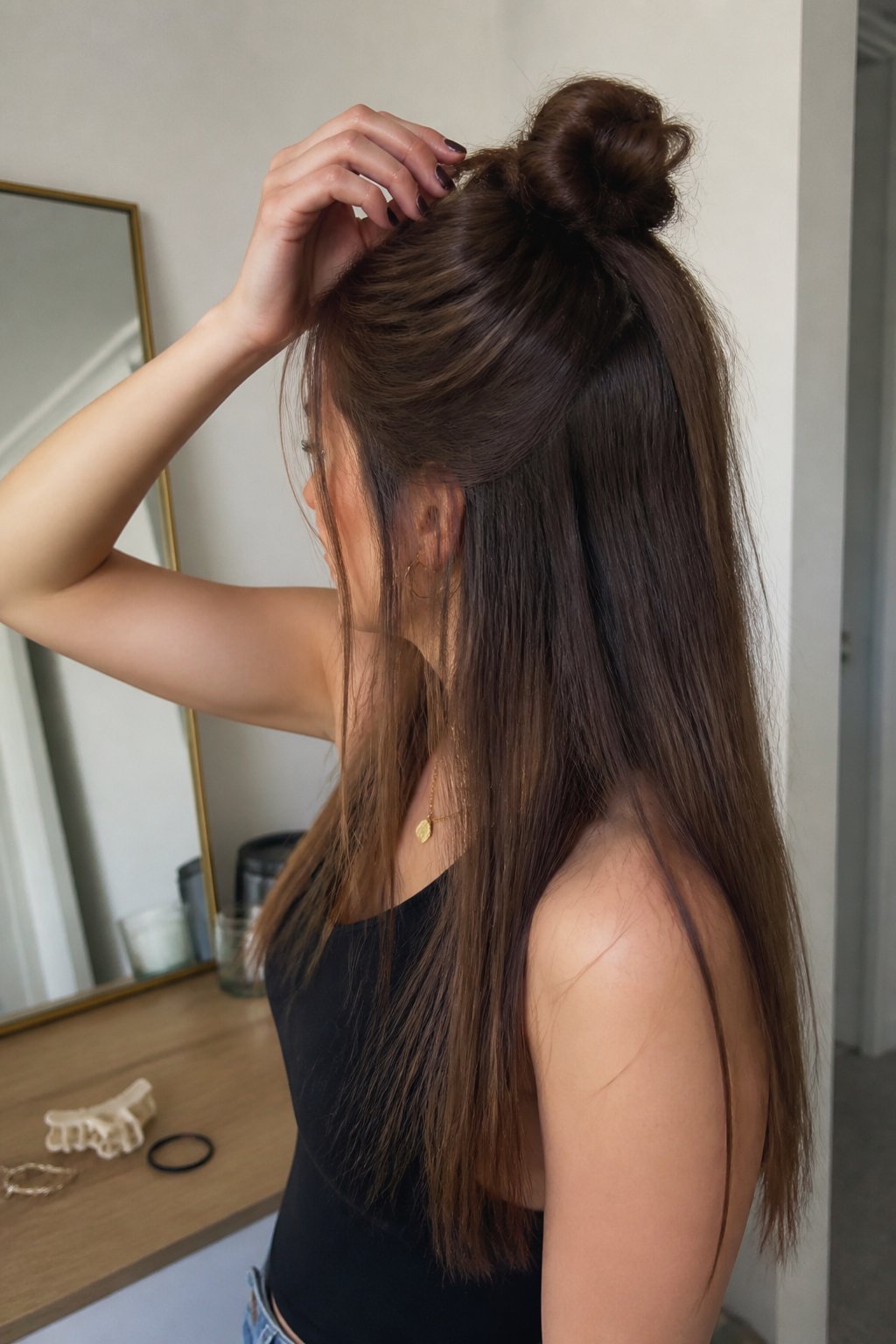A person adjusts a relaxed half-up top knot hairstyle in a softly lit room.