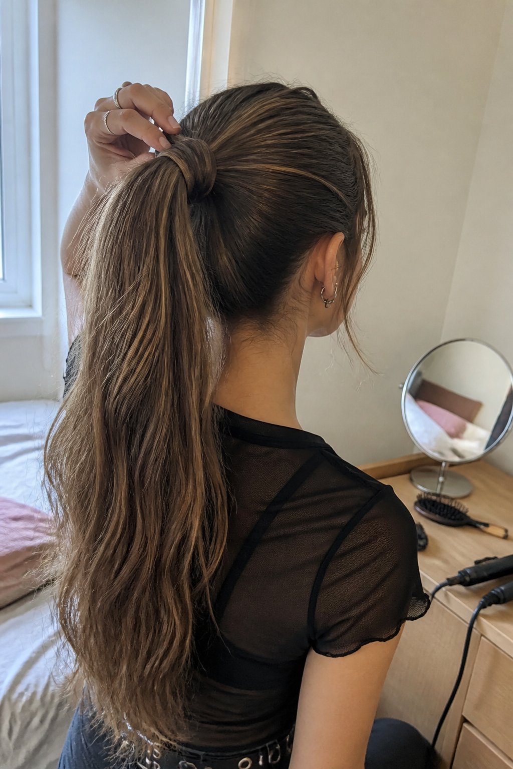 A person adjusts a high ponytail with a section of hair wrapped around the tie while getting ready in a bedroom.