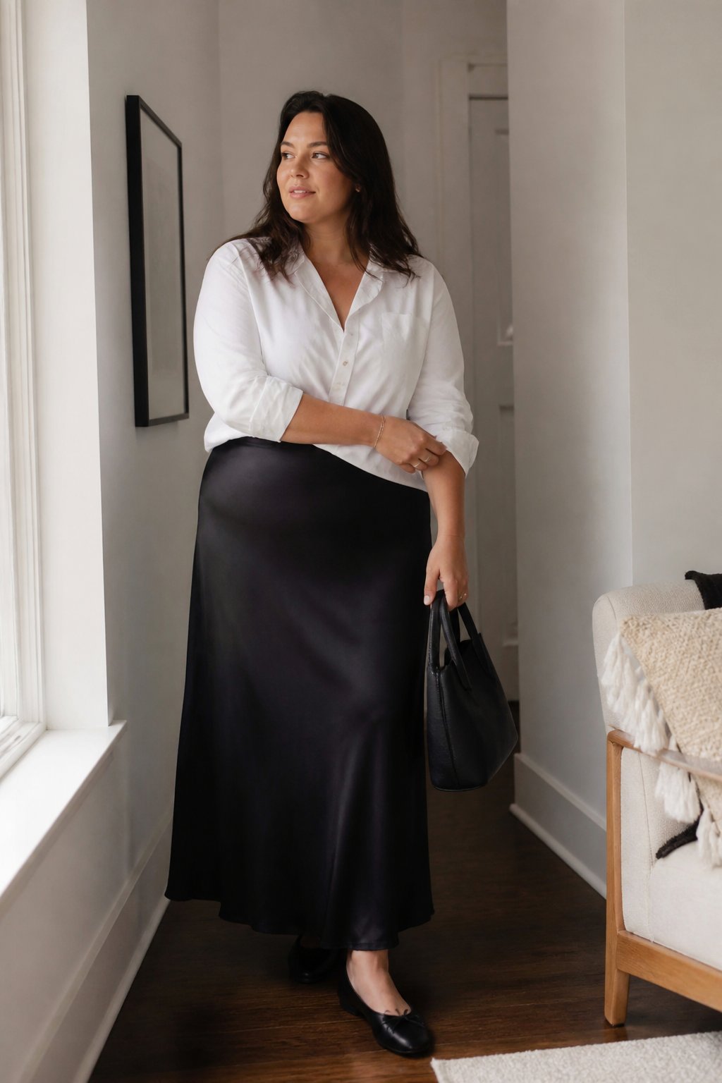A plus-size woman wears a black satin maxi skirt with a white button-down shirt in a softly lit home.