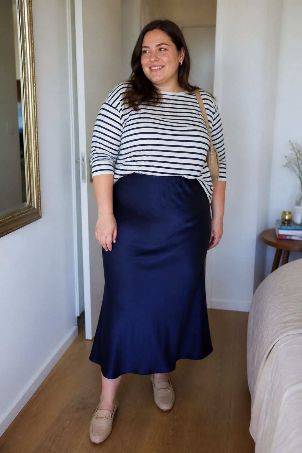 A plus-size woman wearing a navy satin midi skirt and striped Breton top in a softly lit everyday interior.