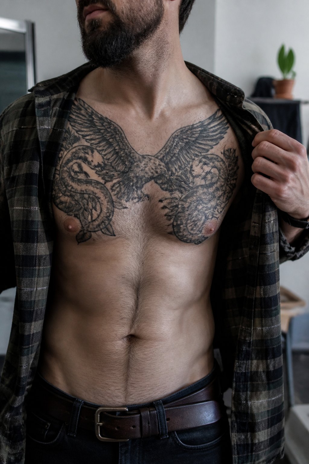 A man with visible chest tattoos stands indoors with his shirt open in soft daylight.