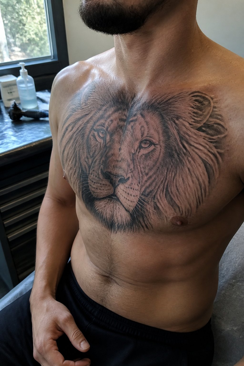 A man displays a realistic lion tattoo across his upper chest in a tattoo studio.