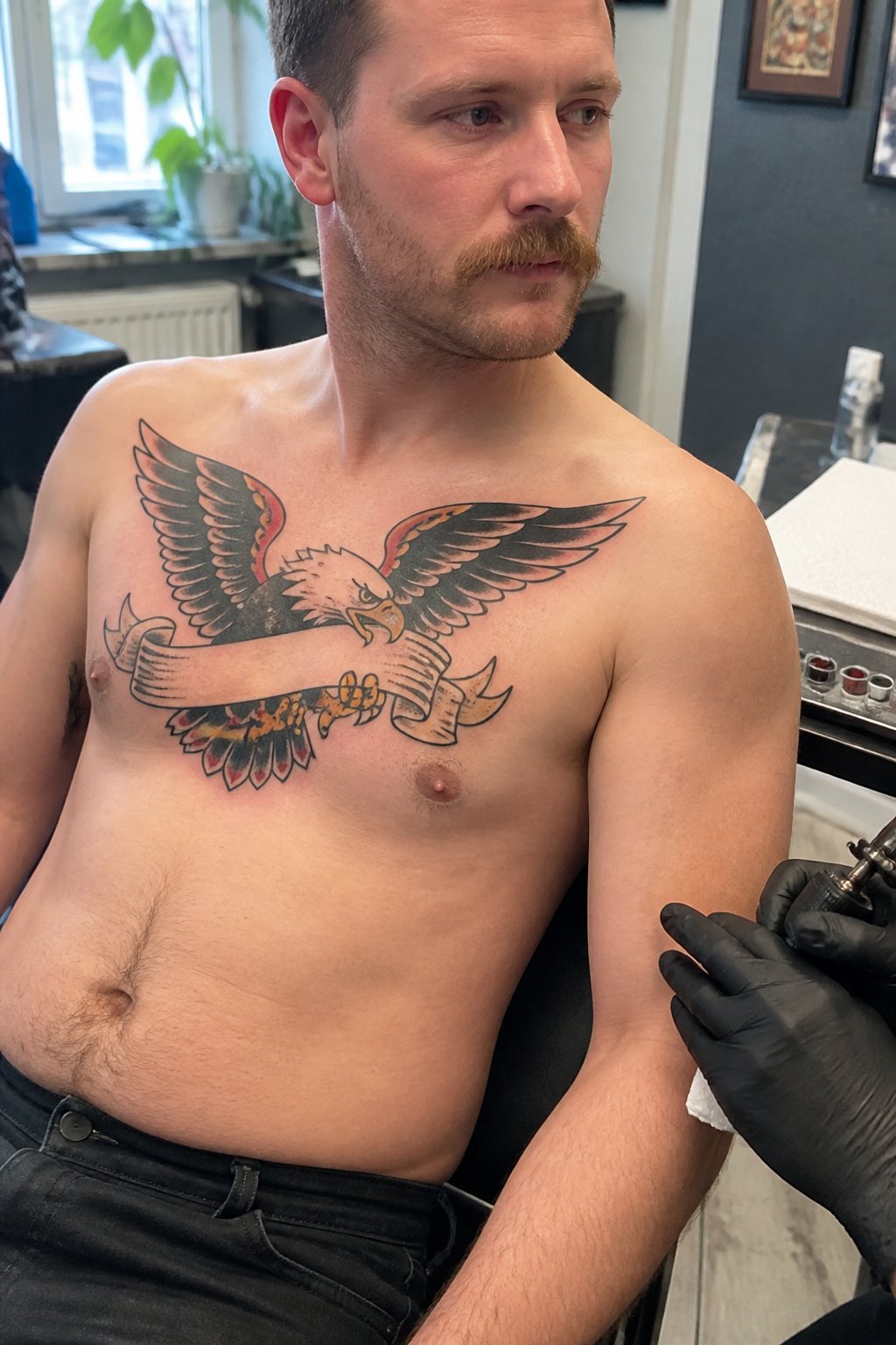 A man sits in a tattoo studio while an artist works on a traditional bald eagle and blank banner tattoo across his chest.