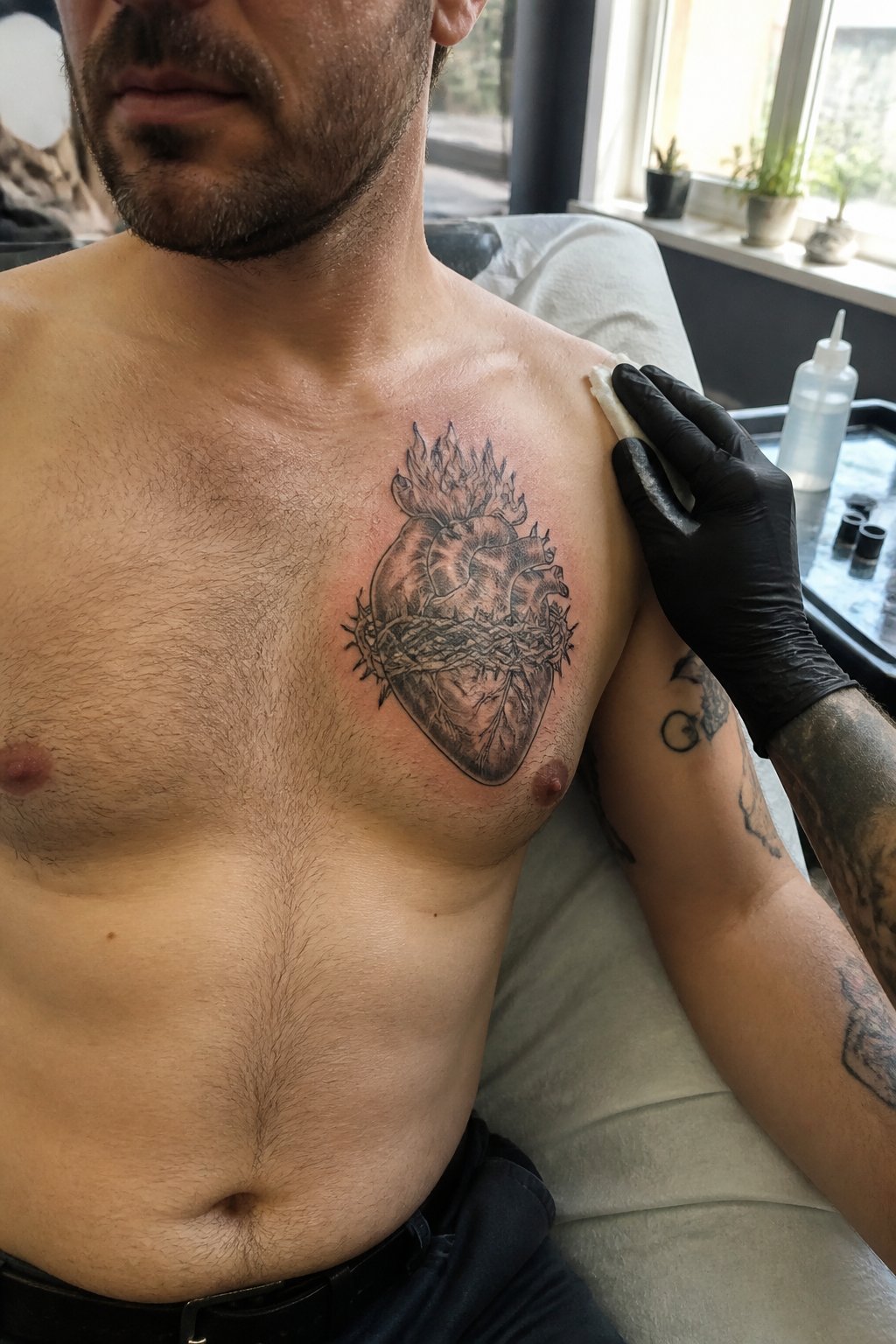 A man displays a sacred heart tattoo on the left side of his chest while a tattoo artist works nearby.