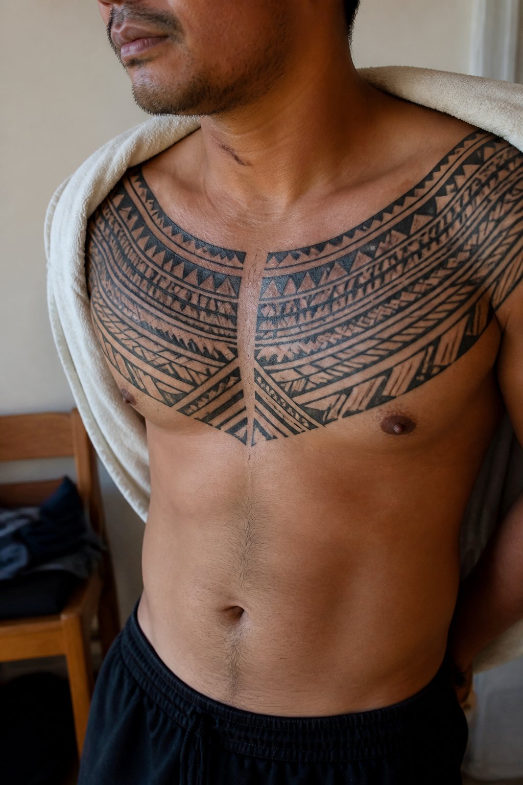 A man displays a bold geometric Polynesian-inspired tattoo across his upper chest in a softly lit indoor setting.