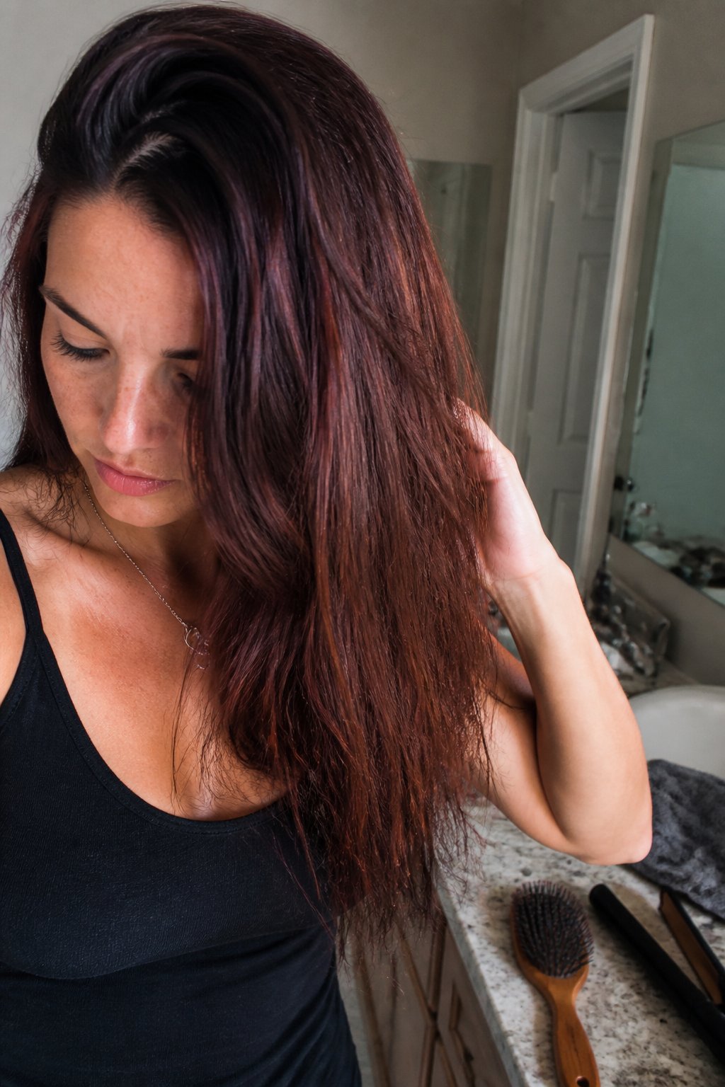 A brunette person with warm mahogany-brown hair stands beside a softly lit window in a simple room.
