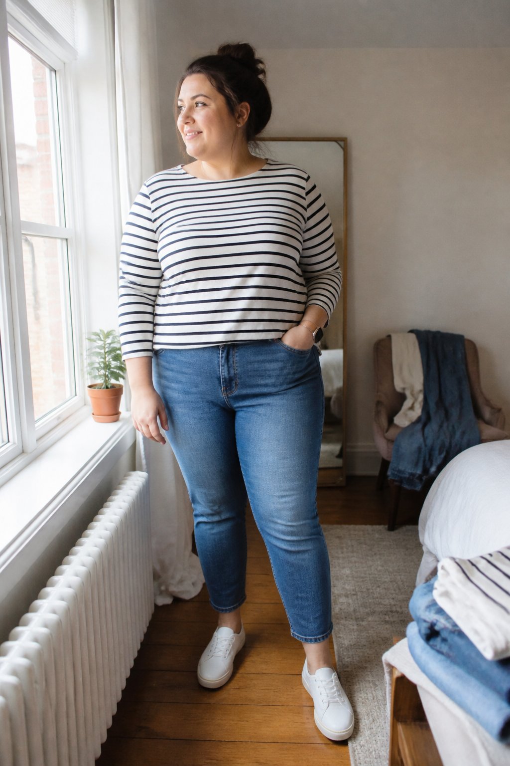 A plus-size woman wearing a striped boat-neck top and ankle-length jeans stands in a softly lit home.